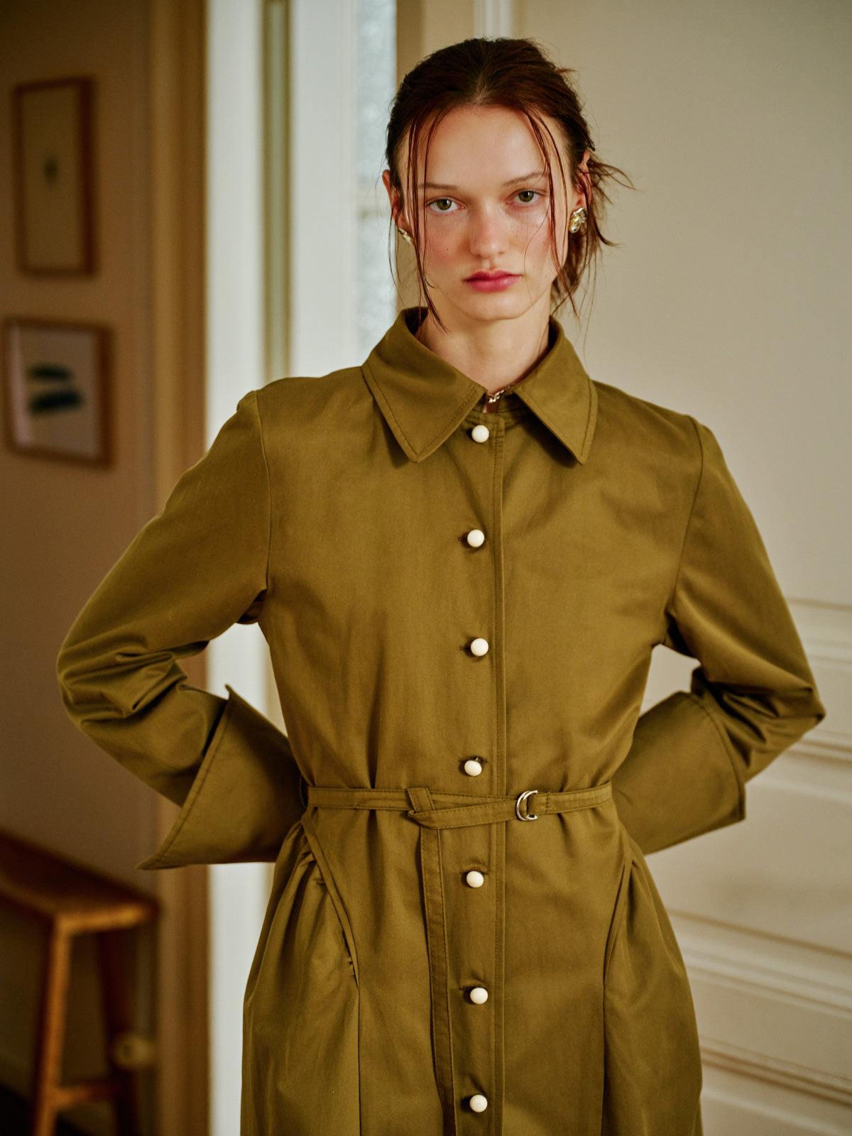 Grotesk Dreams haki trençkot, önden görünümüyle klasik gömlek yaka, sedefli metal düğmeler ve belde kemer detayıyla öne çıkıyor. Detaylara odaklanan bu karede model, minimal bir duruş sergiliyor. WAYT Studio.