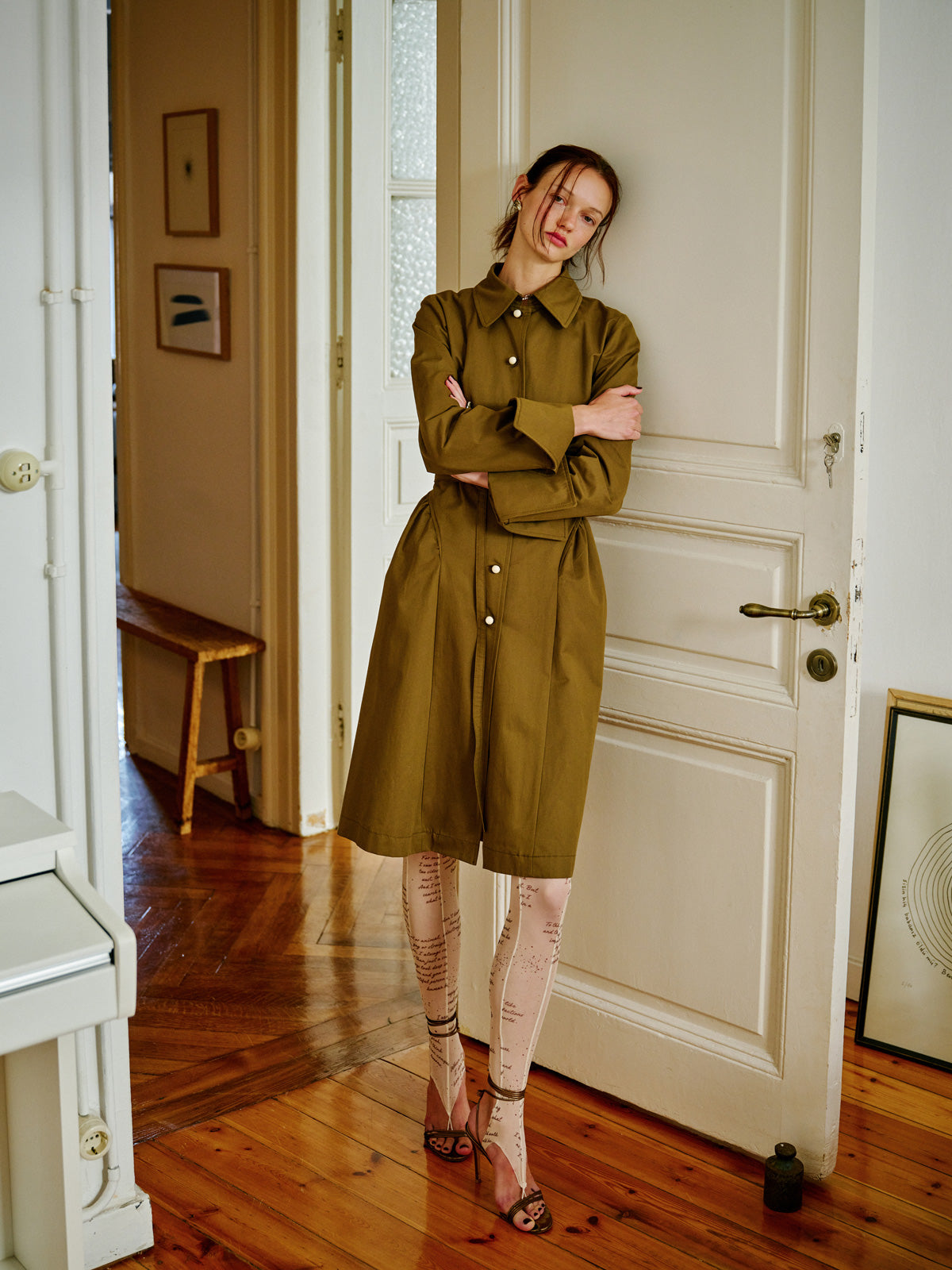 Model, klasik formlu haki renk Grotesk Dreams trençkotu ropuklu sandalet ve yazılı füzo taytla birlikte kapı önünde stilize bir şekilde giyiyor. Zamansız bir görünüm sunan bu tasarım, modern silüetlerle geçmişten gelen formları buluşturuyor. WAYT Studio.