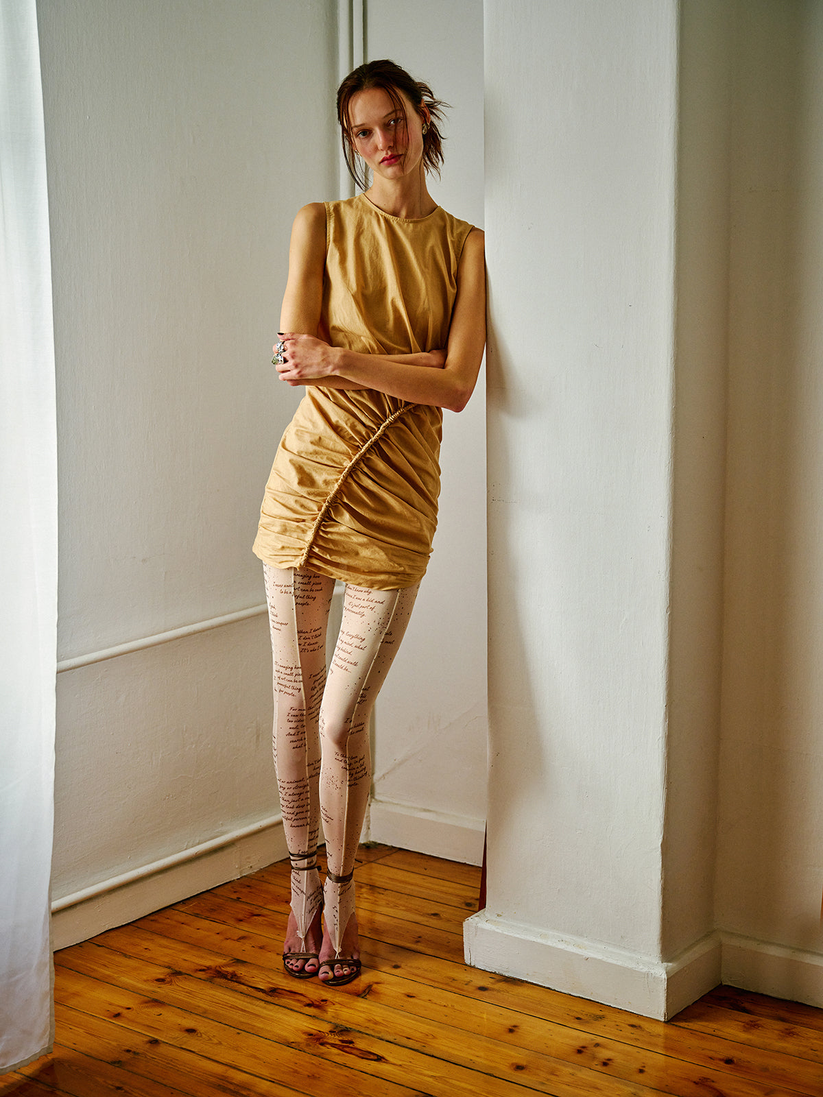 Woman wearing a camel ruched mini dress with asymmetric gathered detailing, styled with printed tights in a softly lit interior.