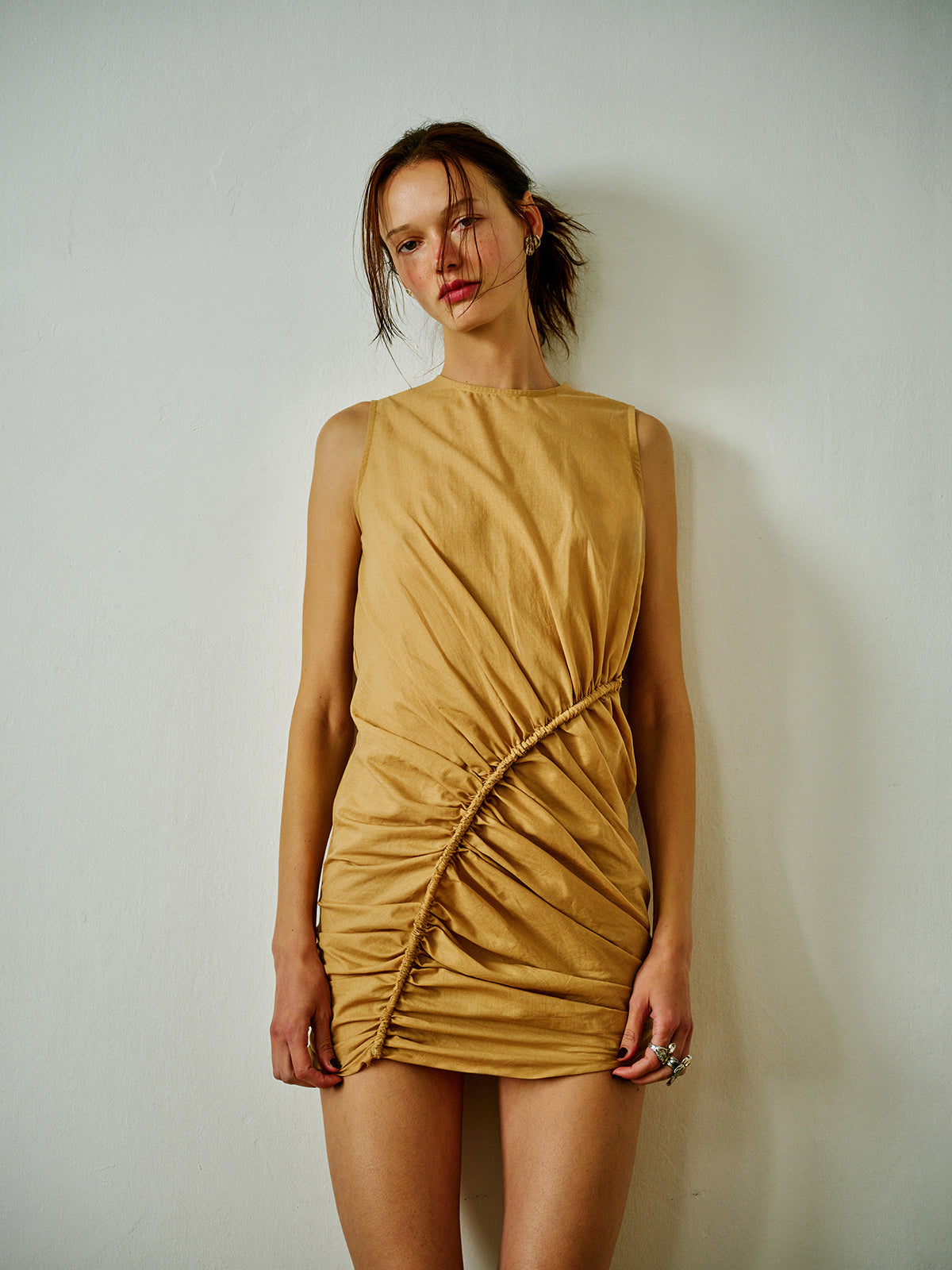 Woman wearing a camel ruched mini dress with asymmetric corded gathering and draped detailing, photographed against a minimal cream wall.