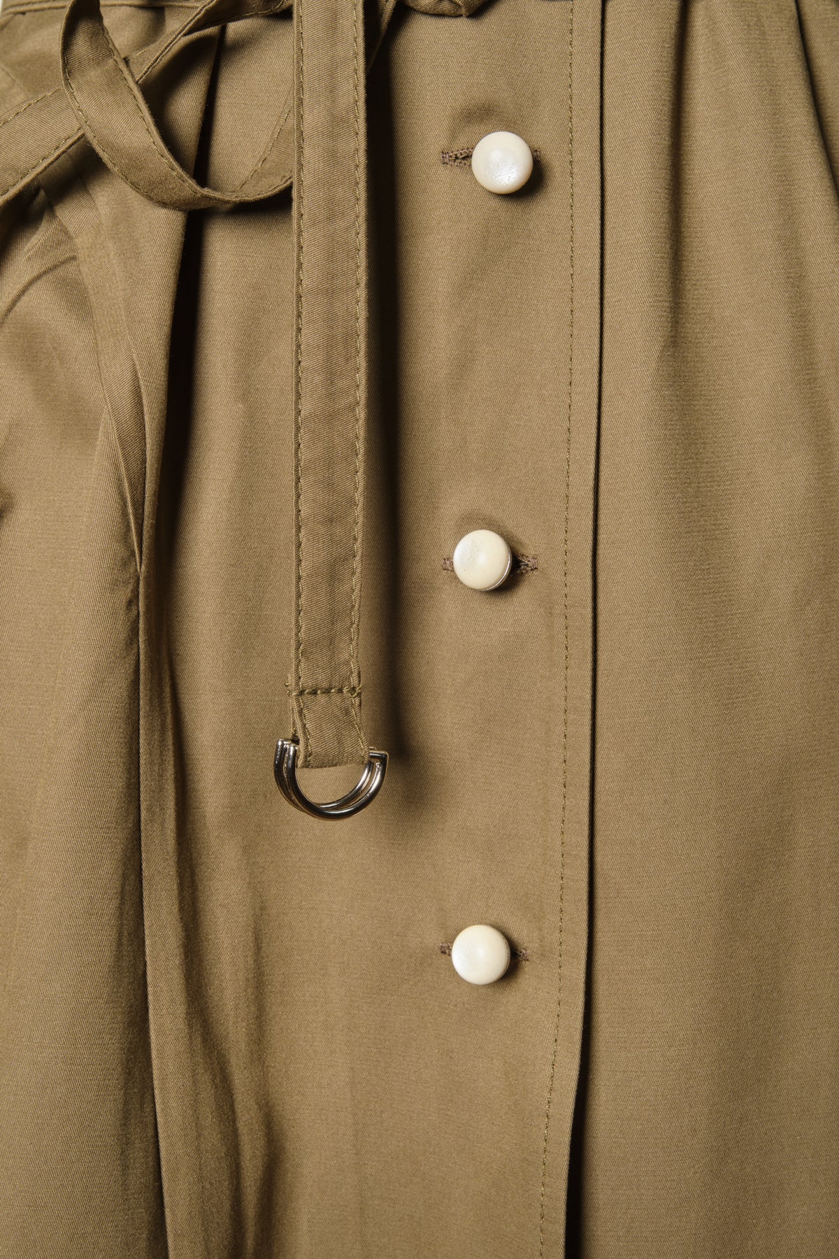 Close-up of WAYT Studio Grotesk Dreams Khaki Trench Coat highlighting the metal D-ring belt and cream enamel buttons on khaki gabardine fabric.
